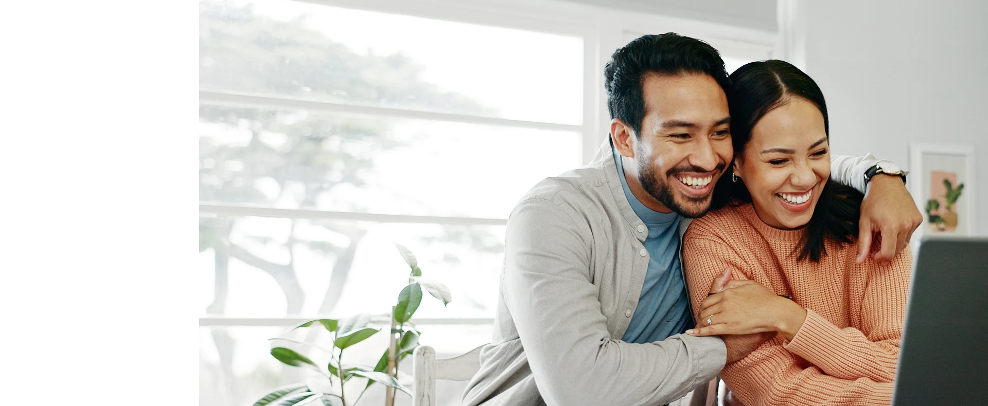 A couple sits close together at a table with laptops and a plant, sharing a cozy moment in a bright, modern workspace.